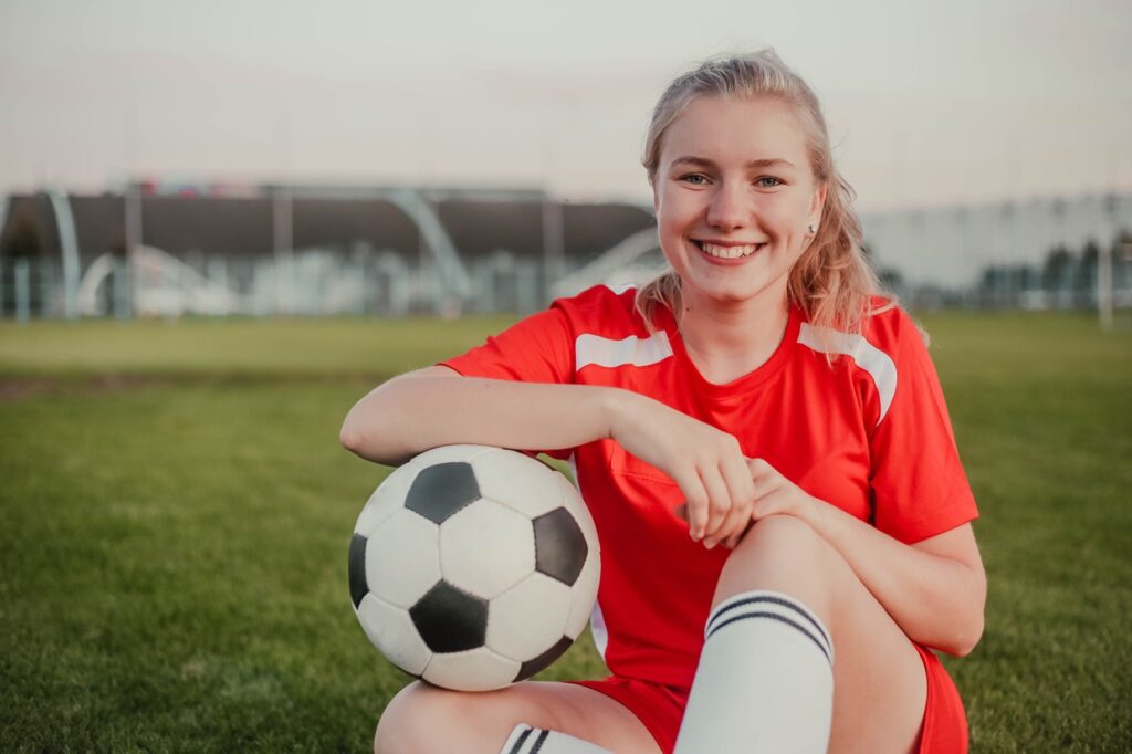 Portret uśmiechającej się dziewczyny grającej w piłkę nożną, siedzącej na boisku razem, trzymającej piłkę.