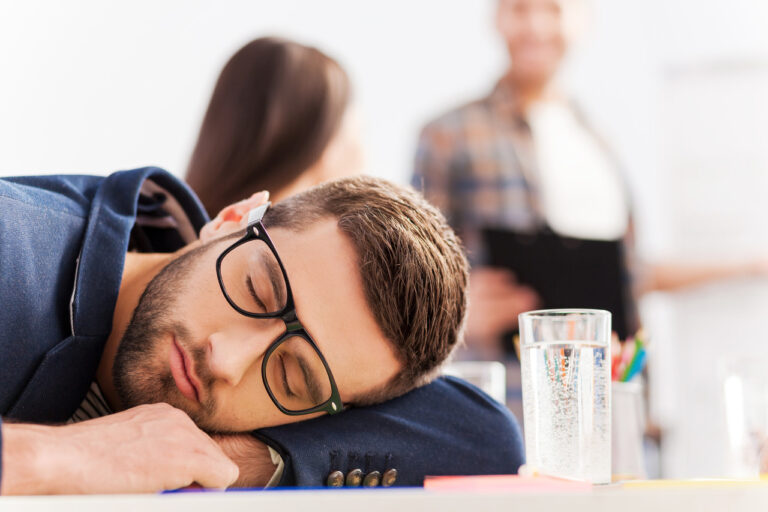 Zmęczony mężczyzna w okularach śpiący na biurku – ilustracja przedstawiająca jak zespół chronicznego zmęczenia może wpływać na codzienne funkcjonowanie.