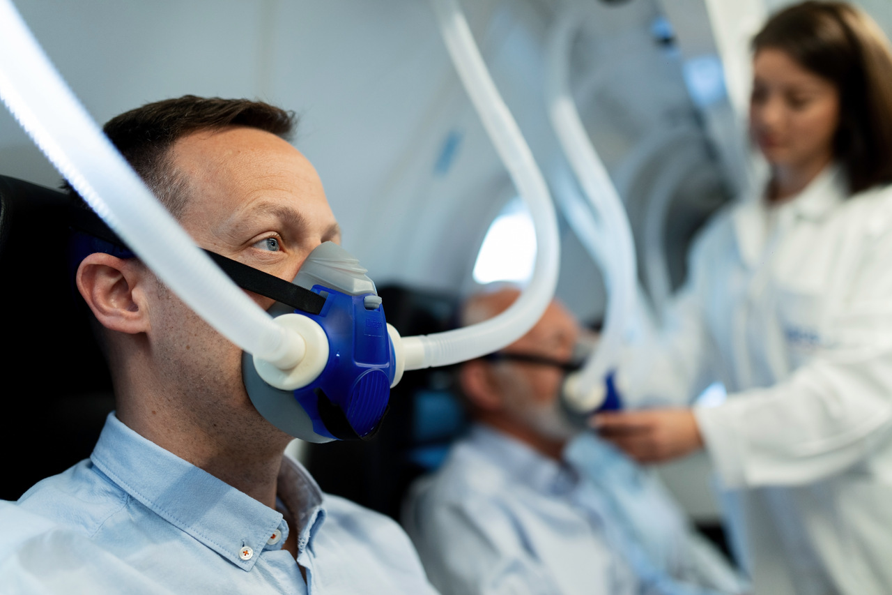 Dorosły mężczyzna z założoną maską do inhalacji, w tle kobieta przeprowadzająca zabieg.
